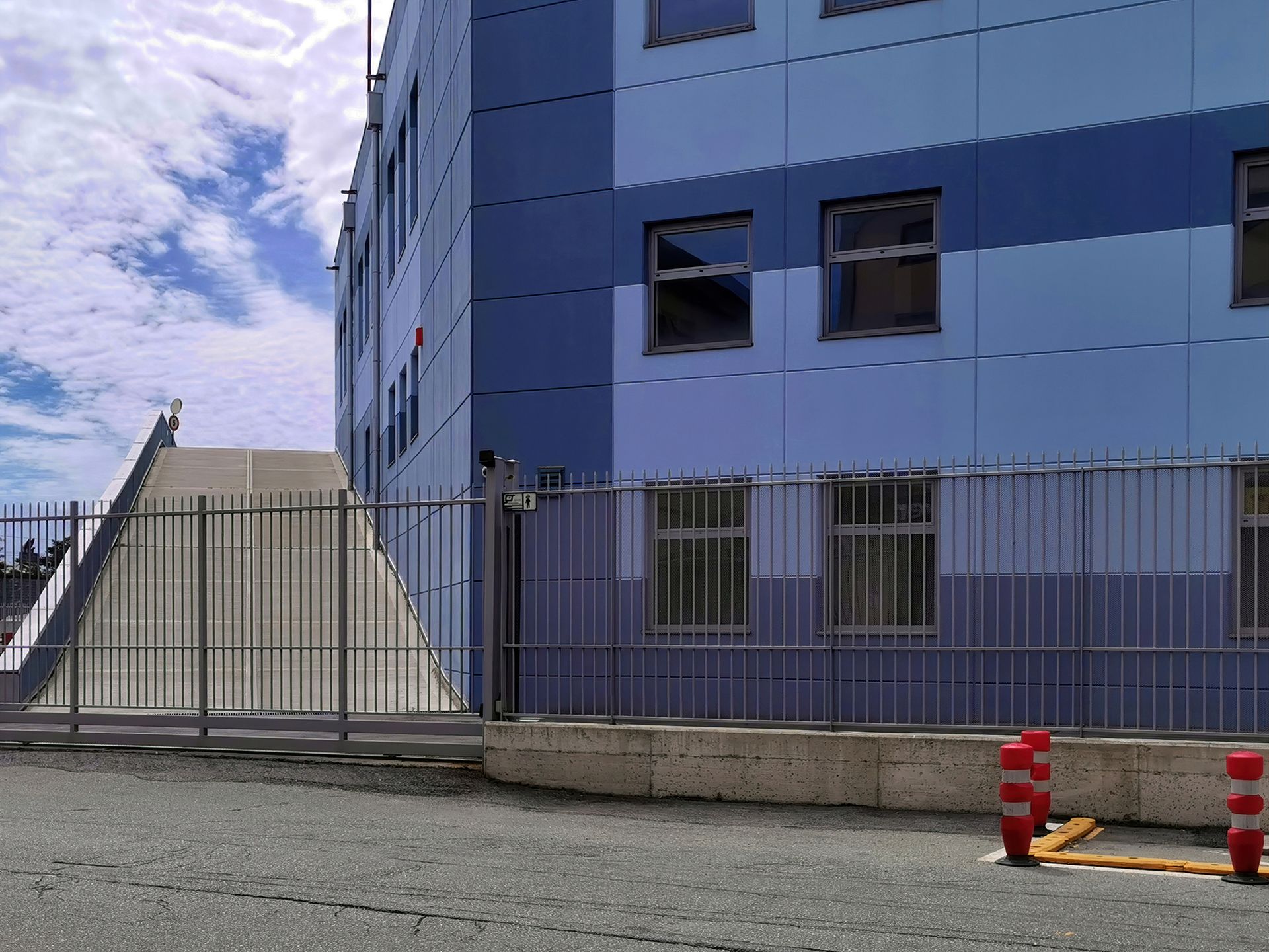 A ramp leading to the entrance of a blue-paneled building, protected by a metal fence and security poles.