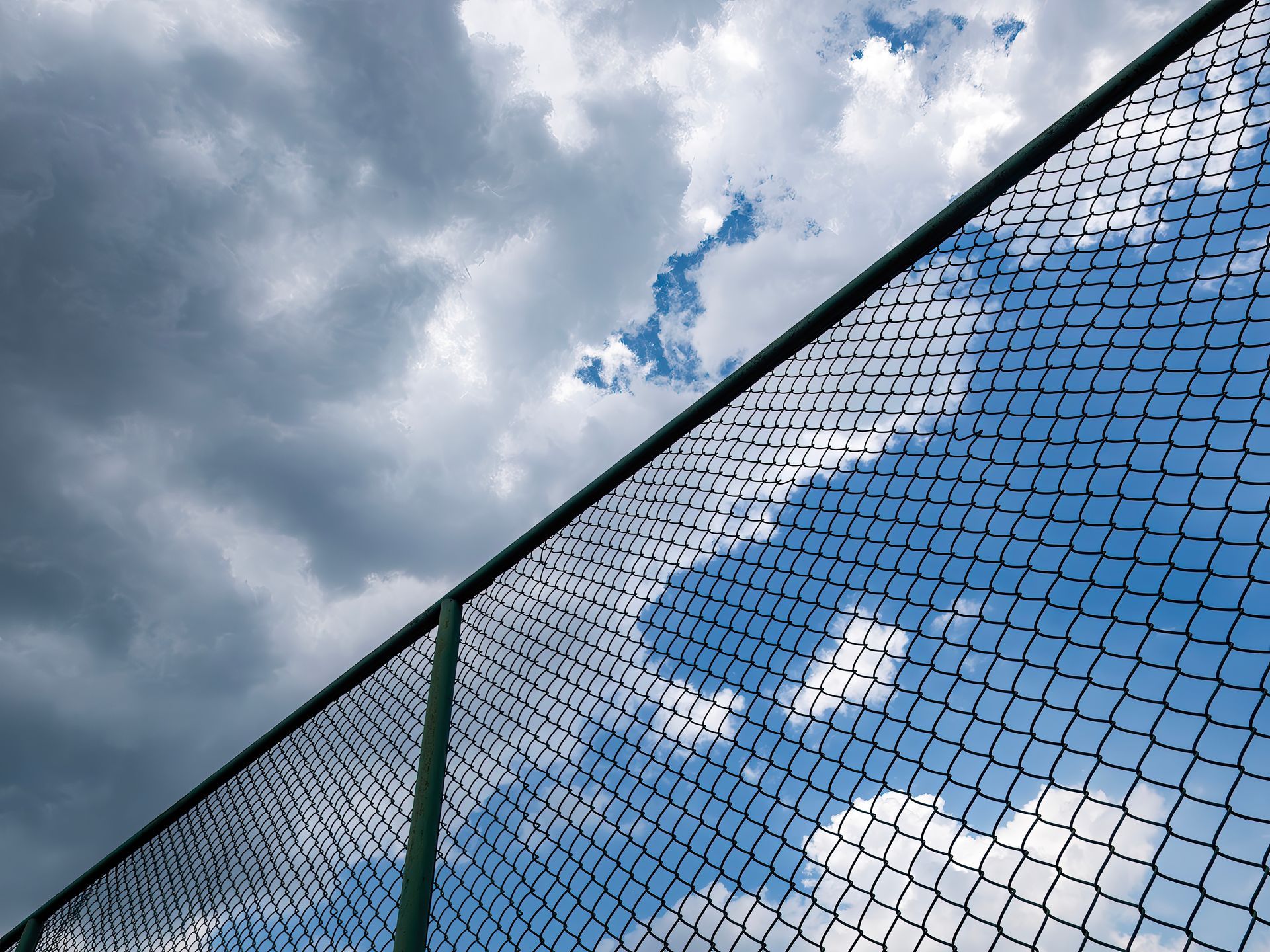 A chain-link fence angles across a frame showing a dramatic sky with both dark, heavy clouds and patches of blue.