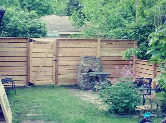 Wooden fence with gate in a backyard, featuring a stone fireplace and table, surrounded by greenery.