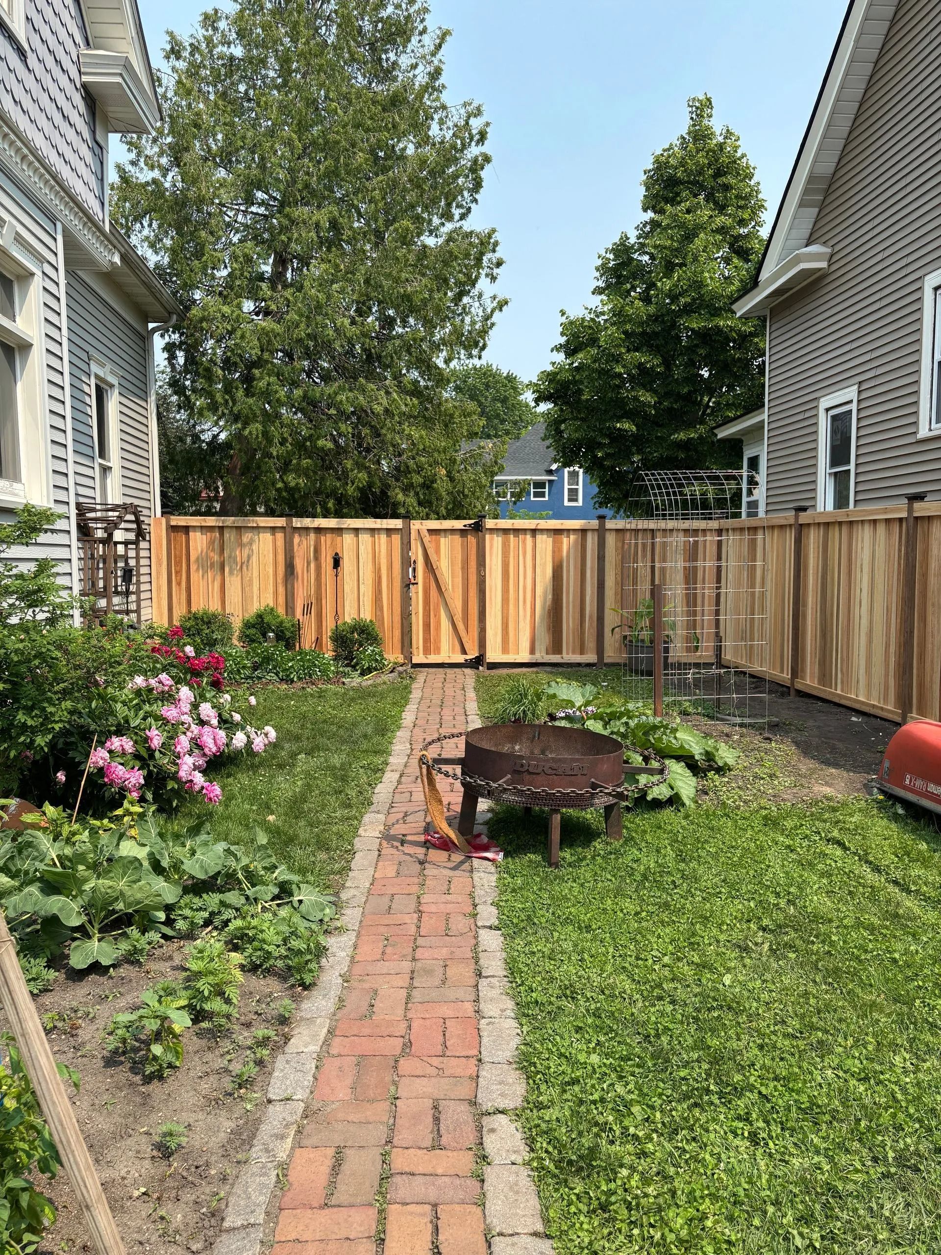 Backyard with a brick path, wooden fence, flowers, fire pit, and houses.