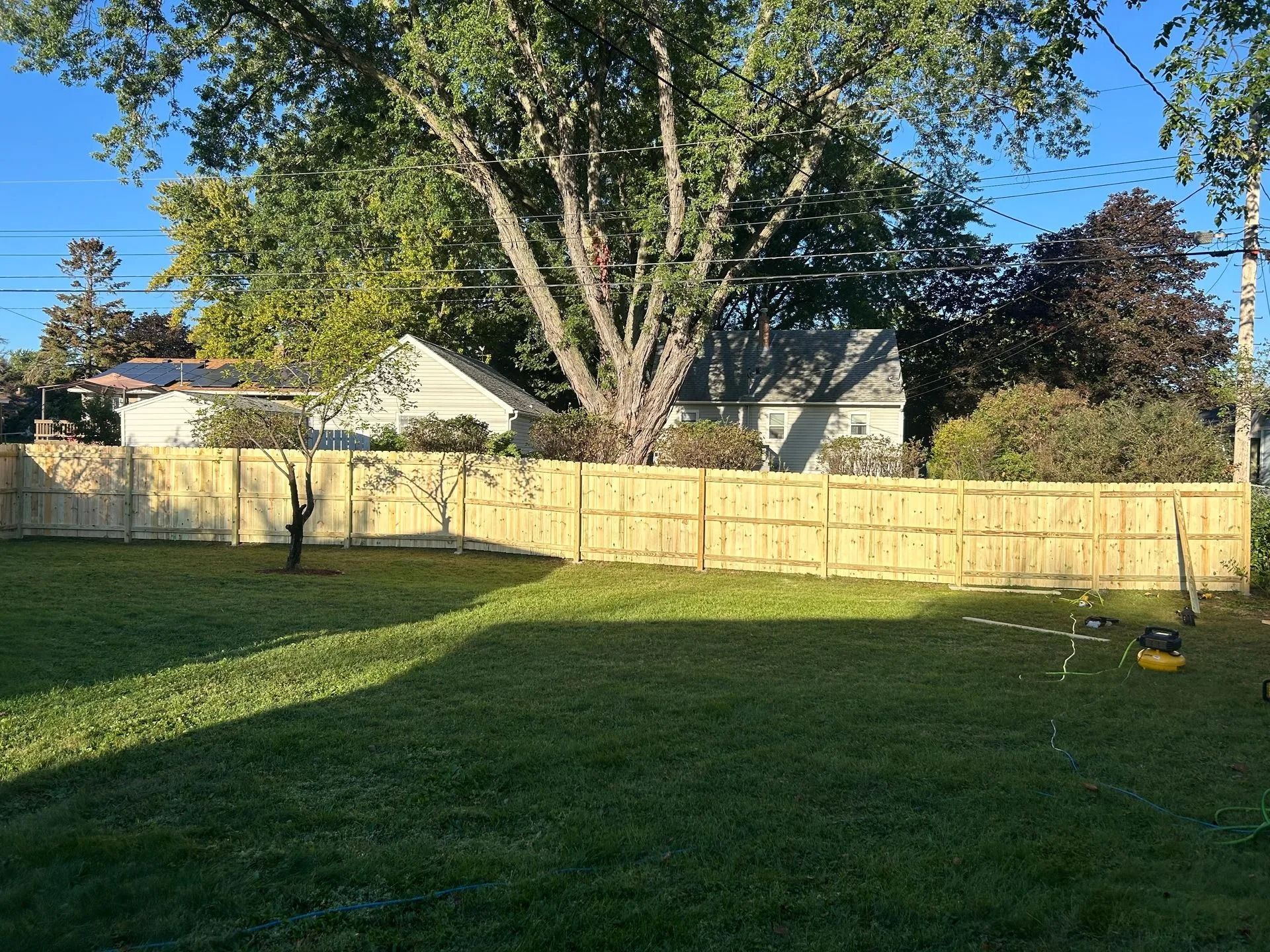A wooden fence surrounds a grassy backyard, with a tree and houses in the background.