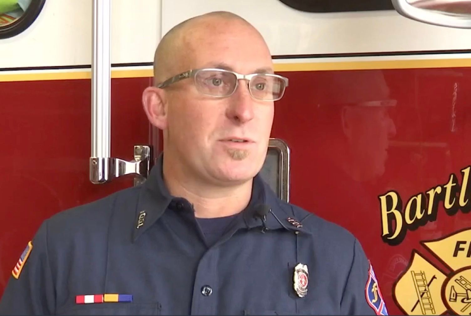 A bald man wearing glasses is standing in front of a fire truck.