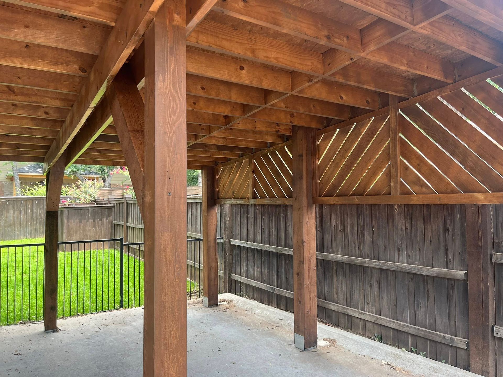 Covered patio with brown wood beams and fence. Green grass and a black fence are in the background.