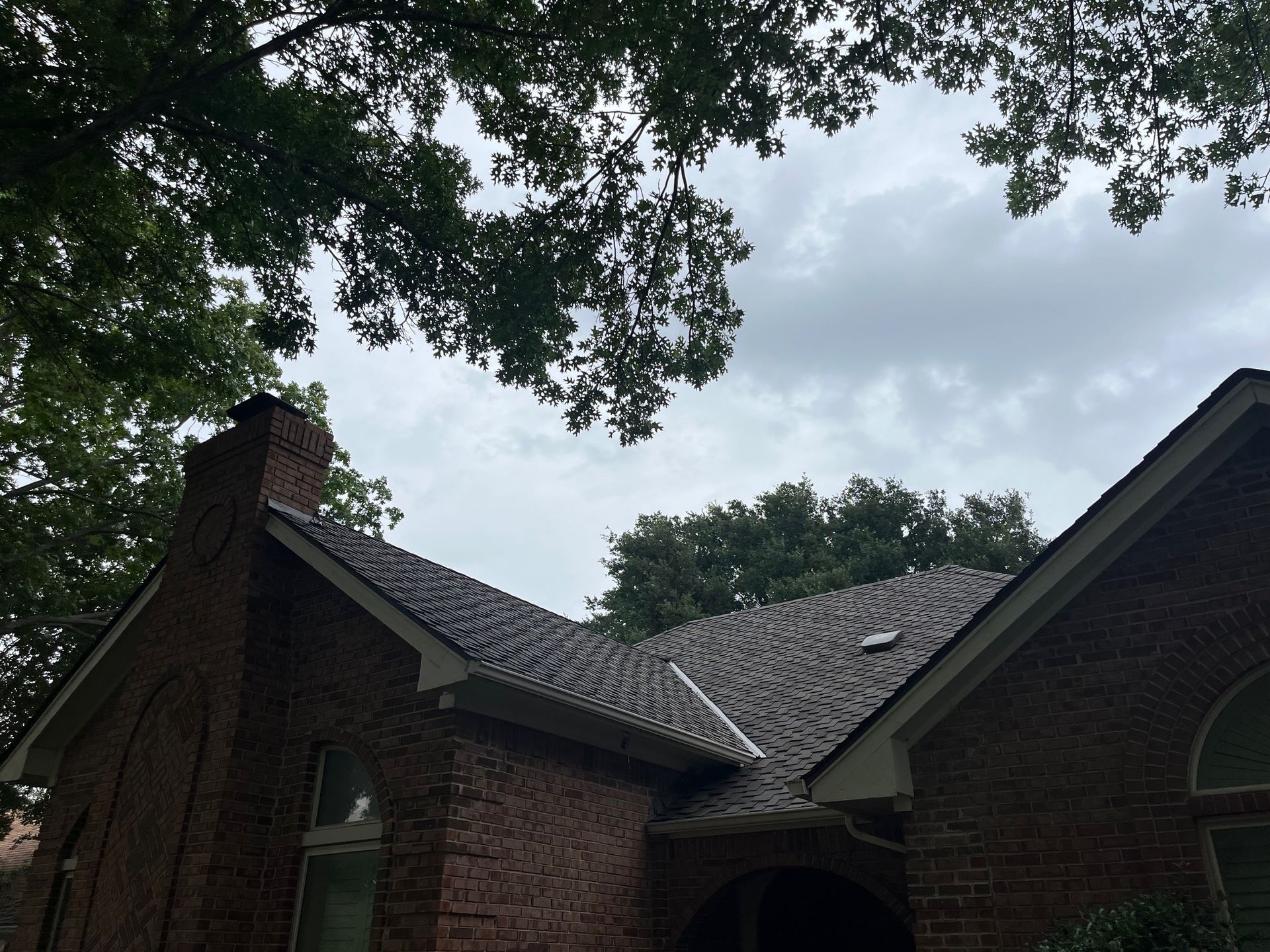 Brick house roof with a chimney under a cloudy sky, framed by tree leaves.