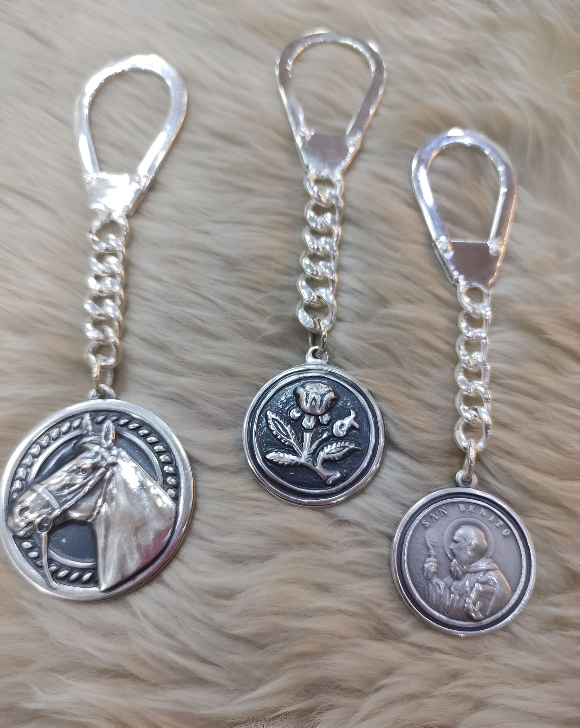 Tres llaveros de plata: cabeza de caballo, flor y medallones de santo unidos a presillas de cadena.