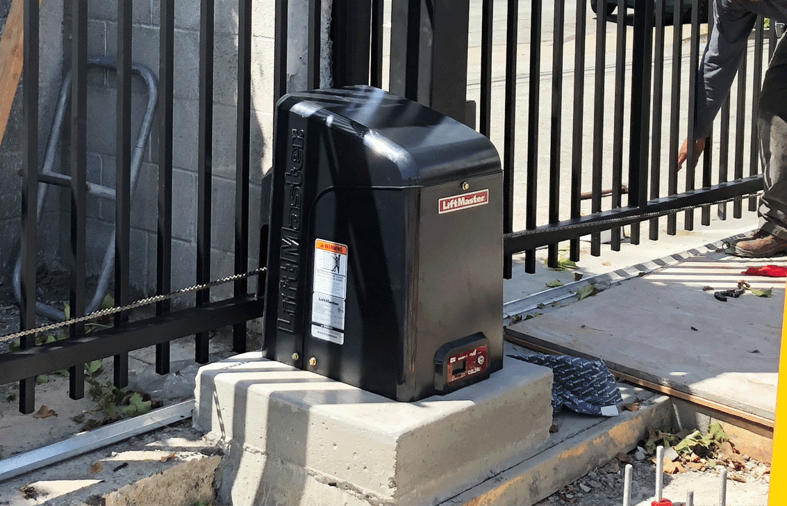 A sliding gate motor is sitting on top of a concrete block next to a fence.
