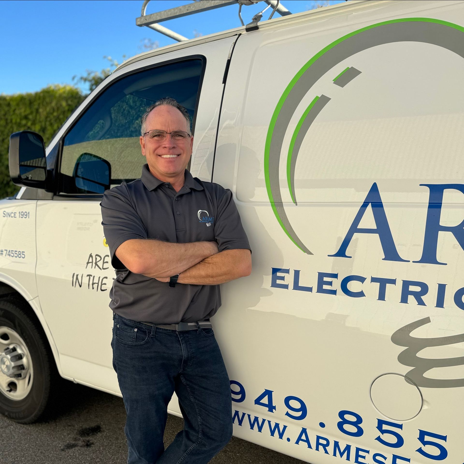 Smiling man standing with arms crossed beside a white electrical service van in sunlight