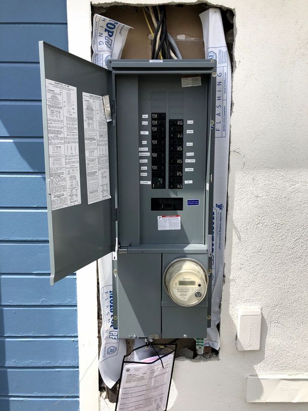 Gray electrical panel with open door, showing breakers. Mounted in a wall, next to a blue-sided building.