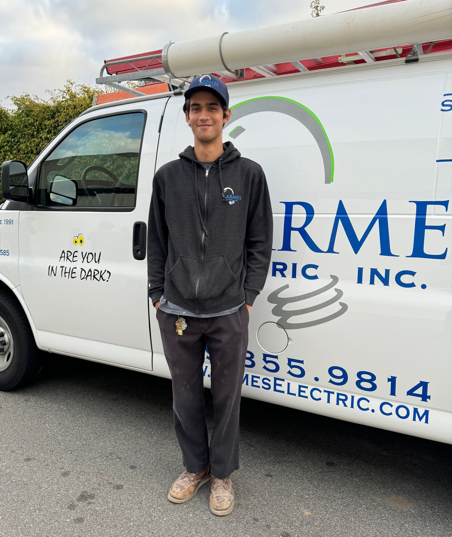 Person standing beside a white service van with blue lettering and a roof ladder, outdoors.