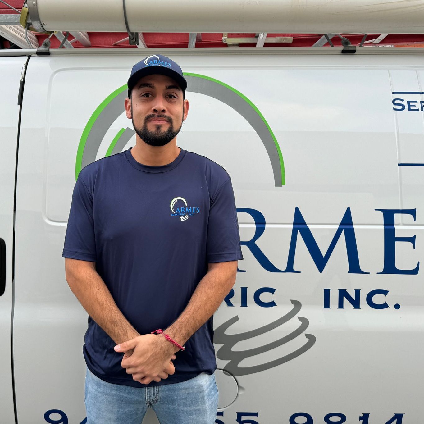 Man standing in front of a service van with company logo, wearing a blue shirt and cap, hands clasped.