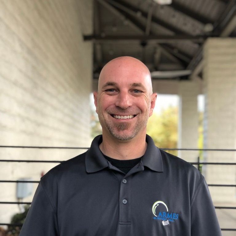 Smiling bald man in a dark polo shirt standing in a covered outdoor corridor.