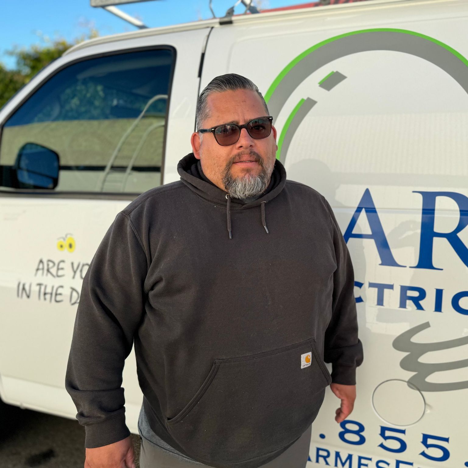 Man in sunglasses and dark hoodie standing beside a white utility truck with company logo.