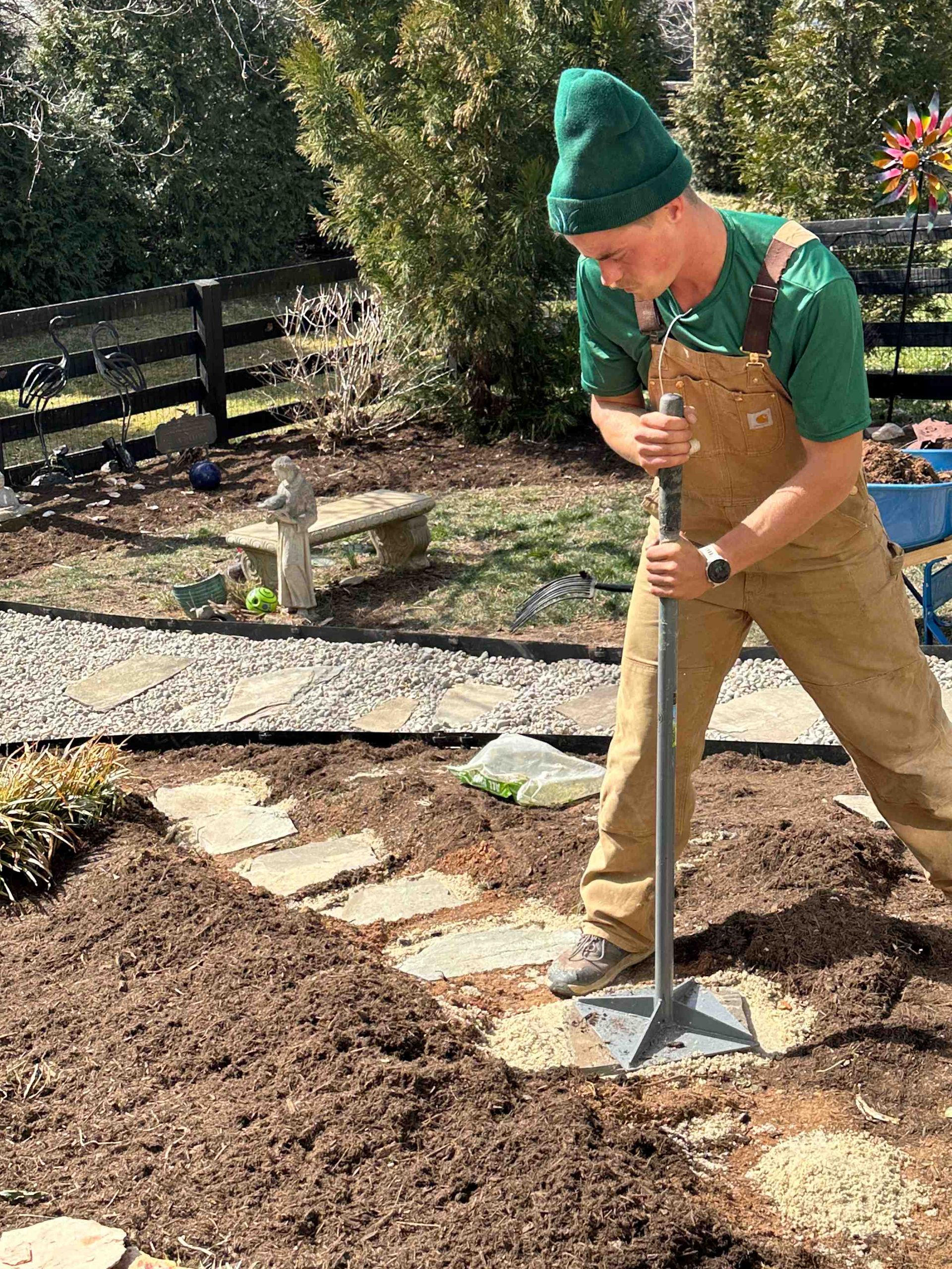 Man in green shirt, overalls, and beanie tamps soil around stone pavers in a garden. Setting is outdoors, sunny day.