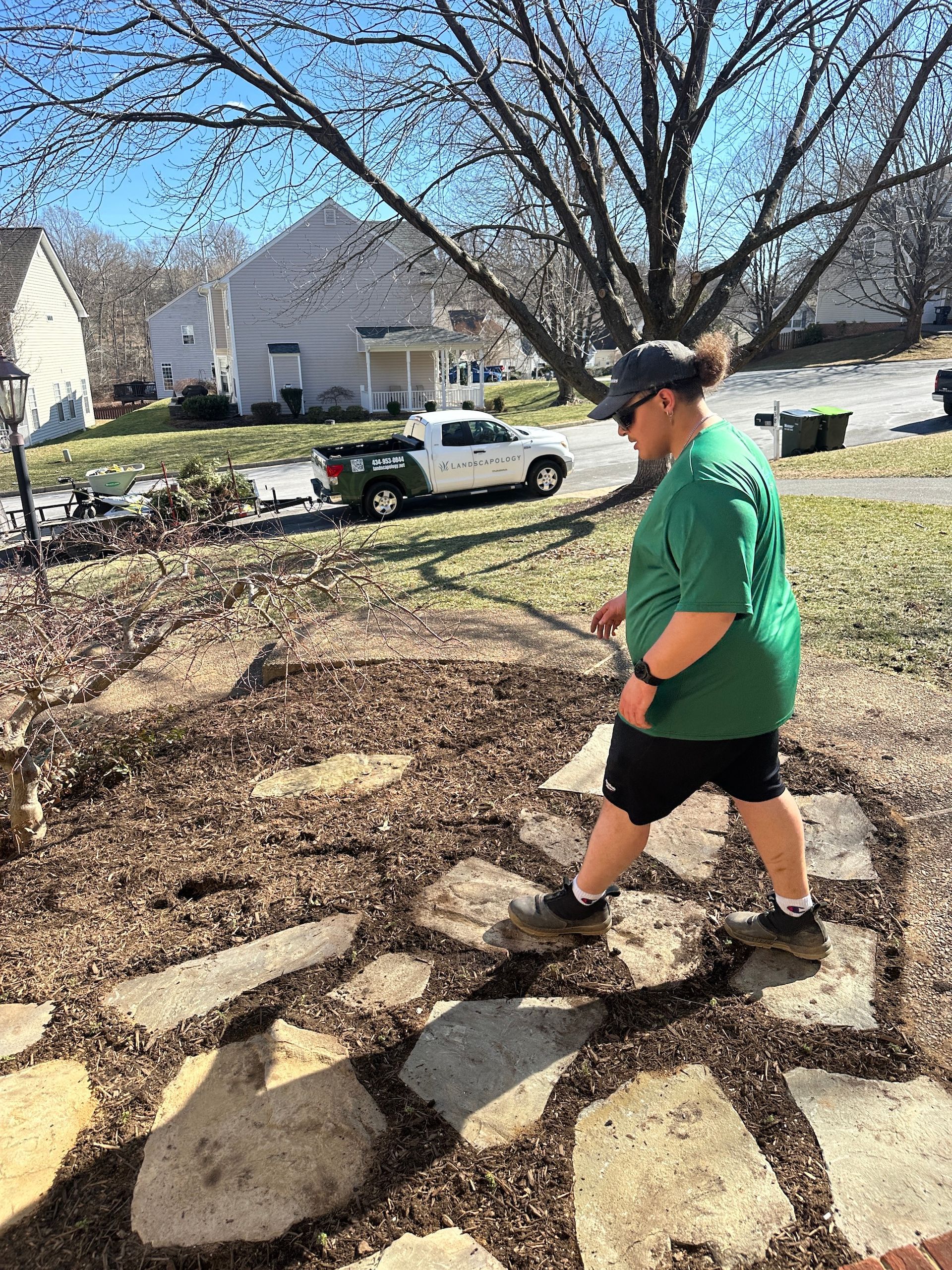 Person in green shirt and black shorts walking on a stone path in a sunny yard with trees and houses nearby