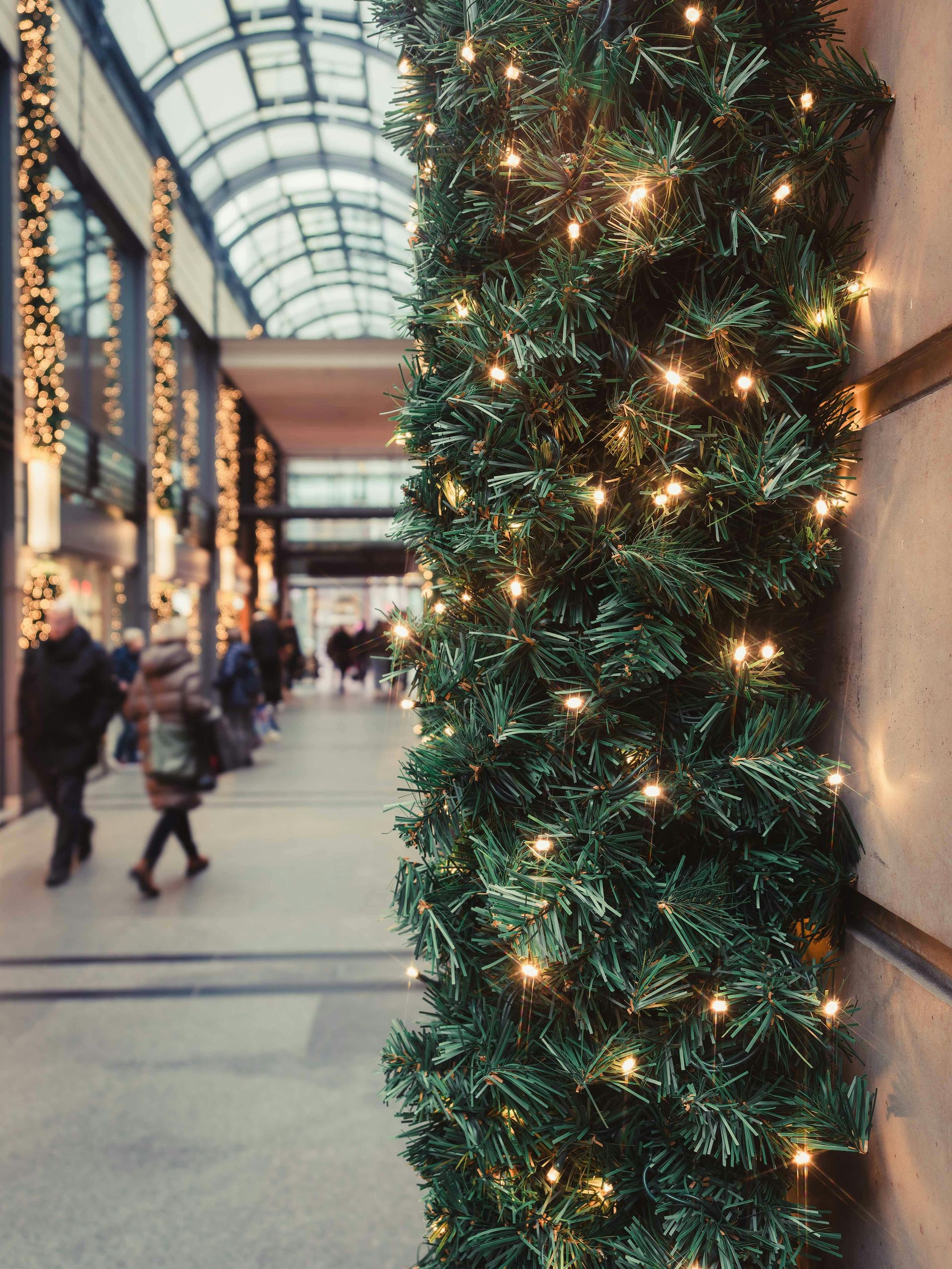 Christmas garland with lights on a pillar in a shopping mall hallway, with people walking.