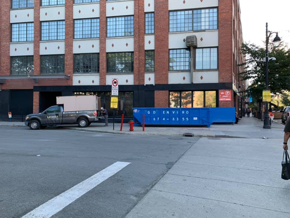 Scène de rue avec un camion, une benne à ordures bleue et un bâtiment en briques à proximité d'un passage piéton