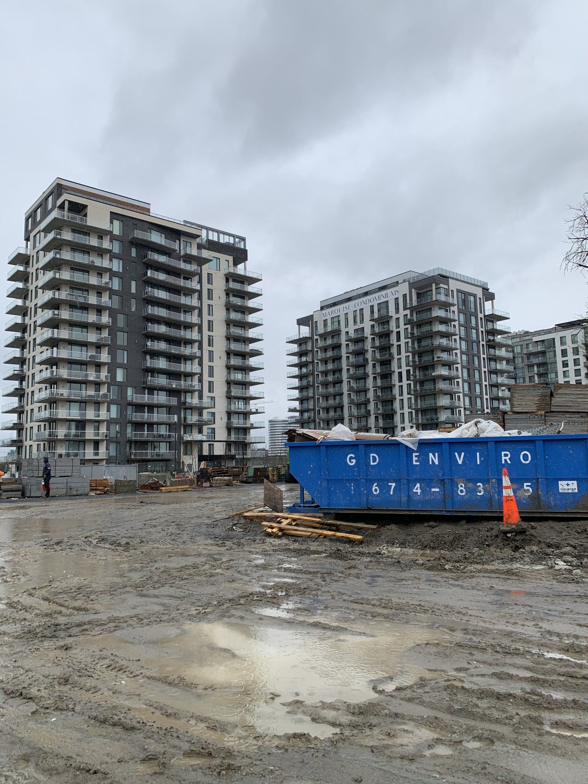 Des immeubles d'habitation se dressent derrière un chantier boueux, avec une benne à ordures bleue et un cône orange sous un ciel gris.
