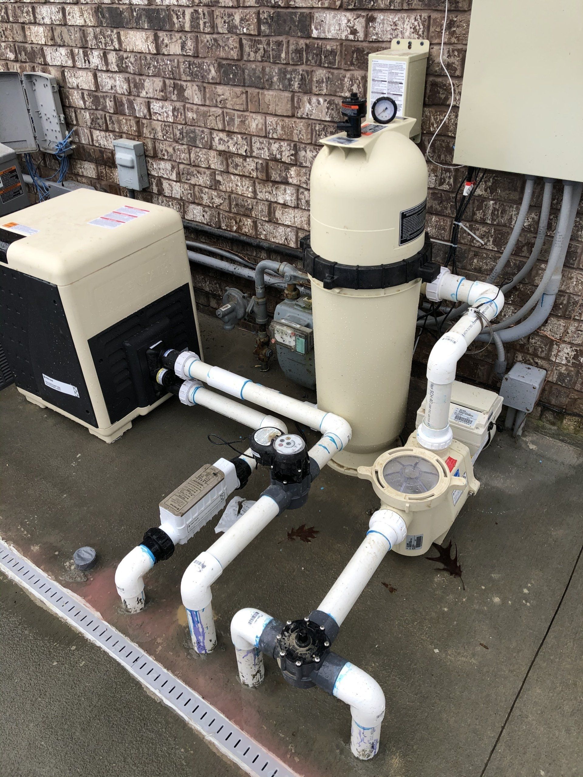 Pool equipment, including a pump, filter, heater, and pipes, against a brick wall.