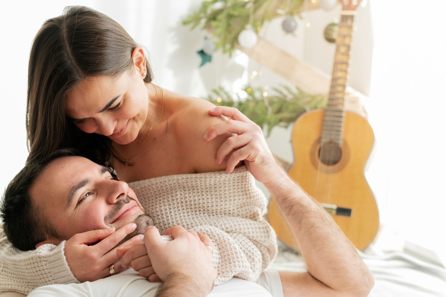 A couple embraces in a bright, cozy room with a guitar in the background, sharing a tender and intimate moment.