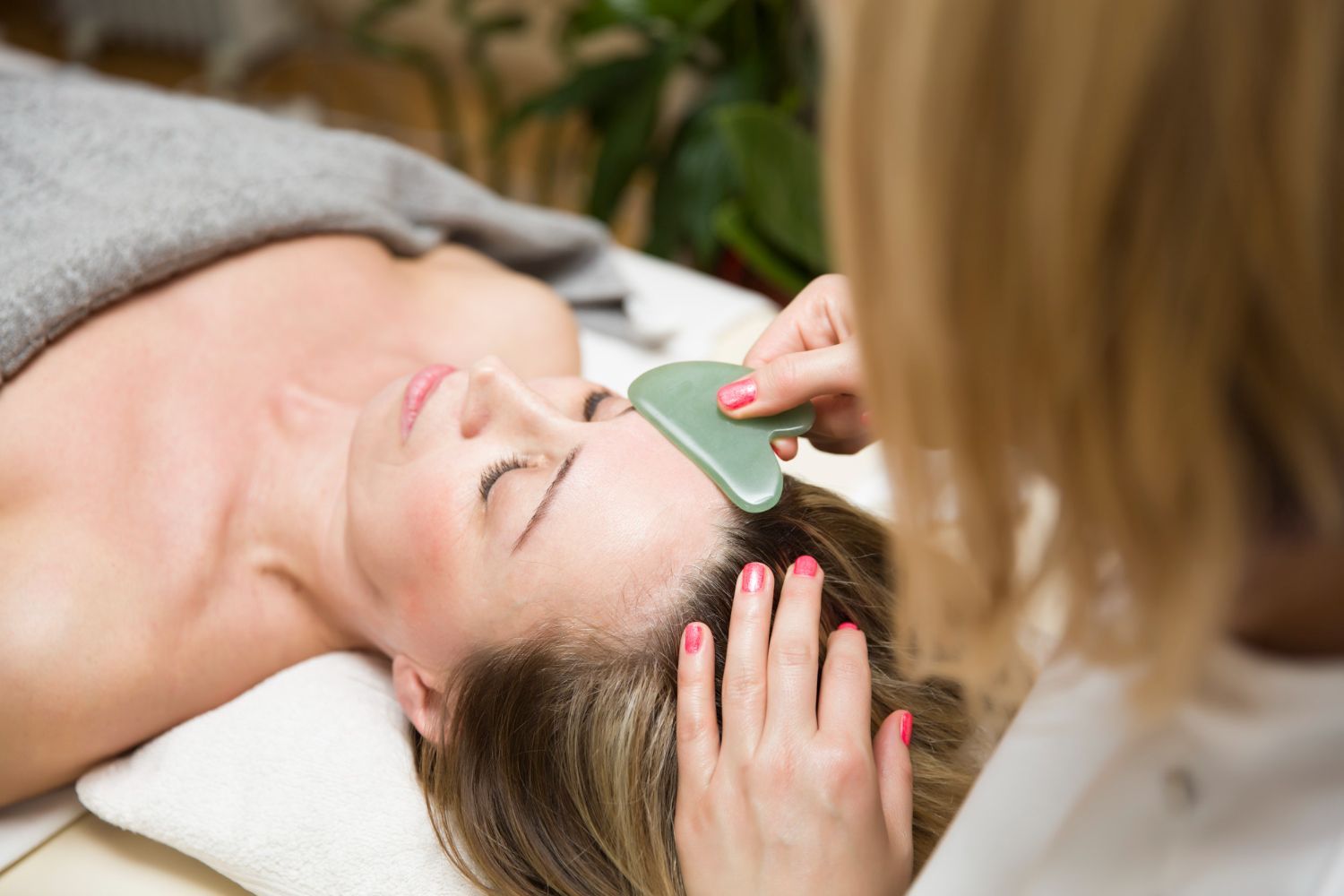 A practitioner uses a green gua sha tool on a person's forehead during a facial treatment.