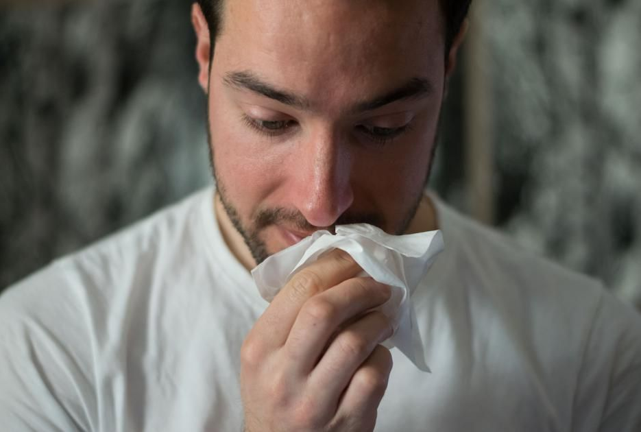 A person in a white shirt holds a tissue to their nose, appearing to be unwell.