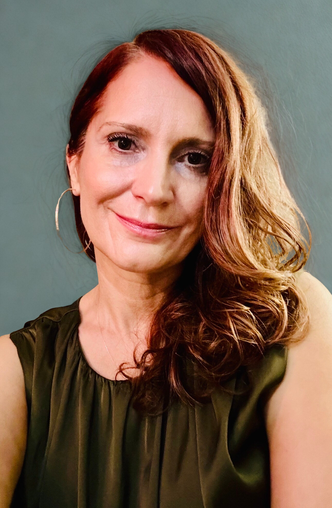 Woman with brown hair smiles, wearing a black top. Lit by sunlight with green plants in background.