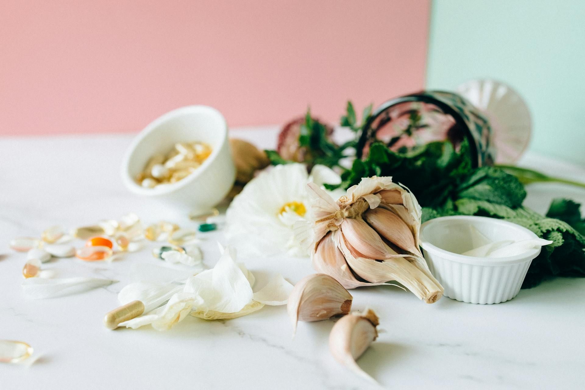 Garlic cloves, white flower petals, and scattered dietary supplements arranged on a white marble surface.