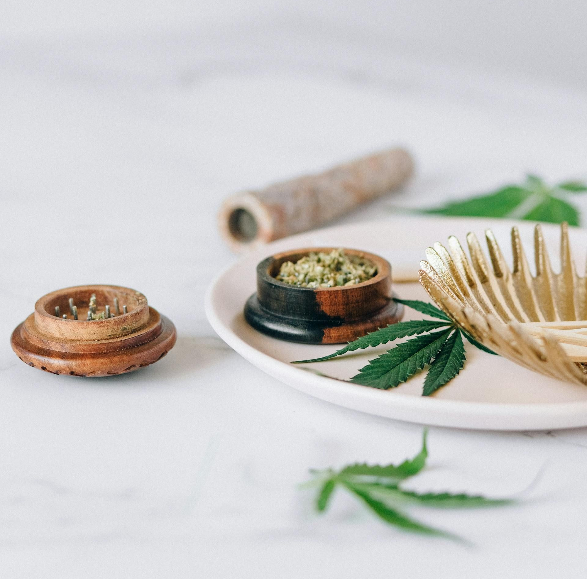 A wooden herb grinder, a small wooden bowl filled with cannabis, and a gold bowl on a white plate with cannabis leaves.
