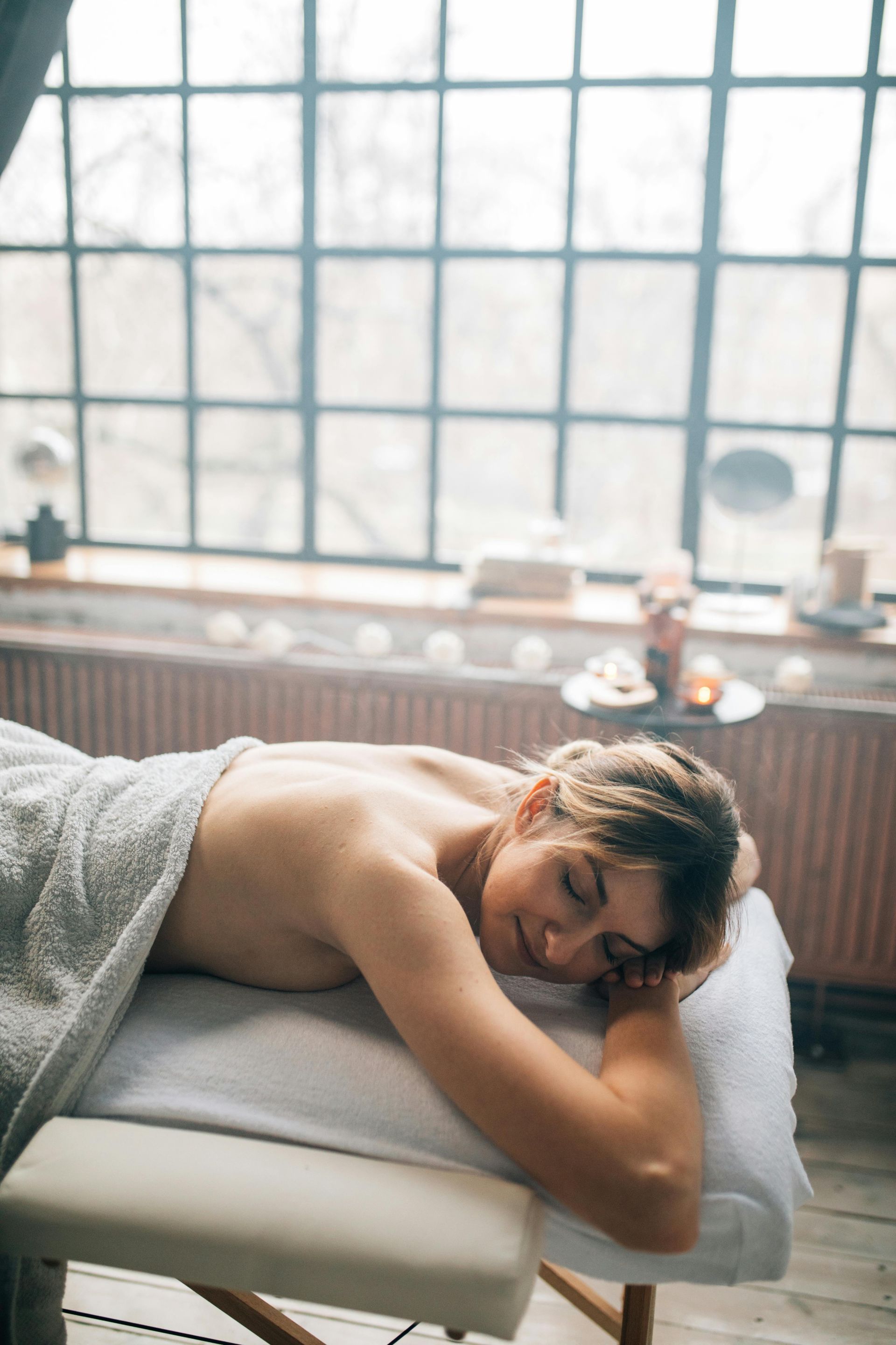 A person rests on a massage table, eyes closed in relaxation, in a room with a large window.