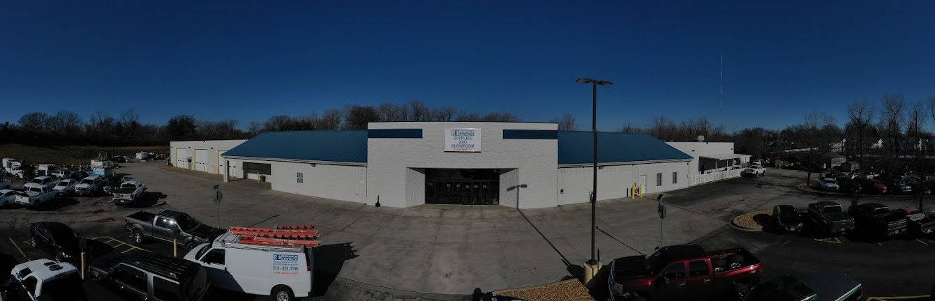 Wide shot of a white building with blue roof under a clear blue sky. Parking lot with vehicles.