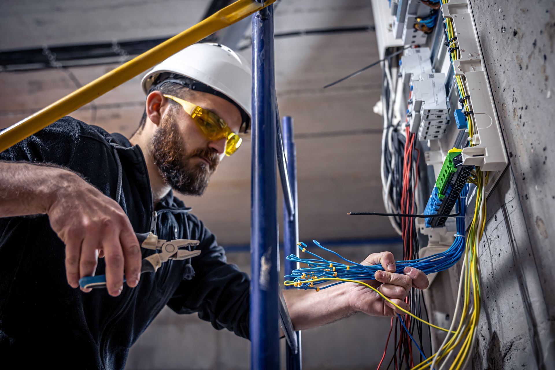 A certified electrician wearing a helmet works in a switchboard with an electrical connecting cable. A certified electrician wearing a helmet works in a switchboard with an electrical connecting cable.