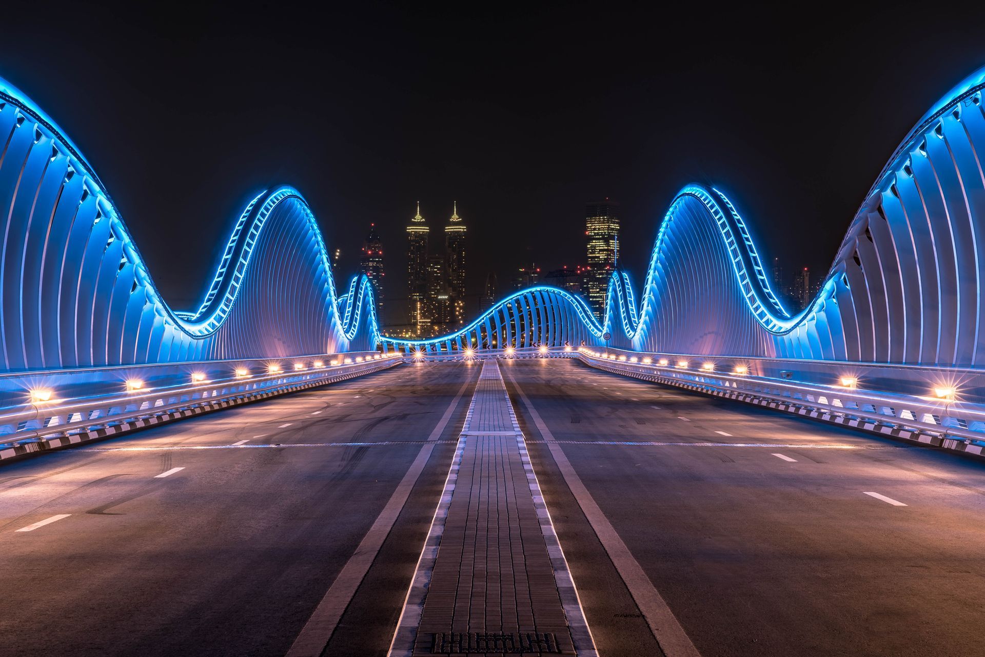 A nighttime view of the Dubai Water Canal Bridge illuminated in blue, with city skyline in the background.