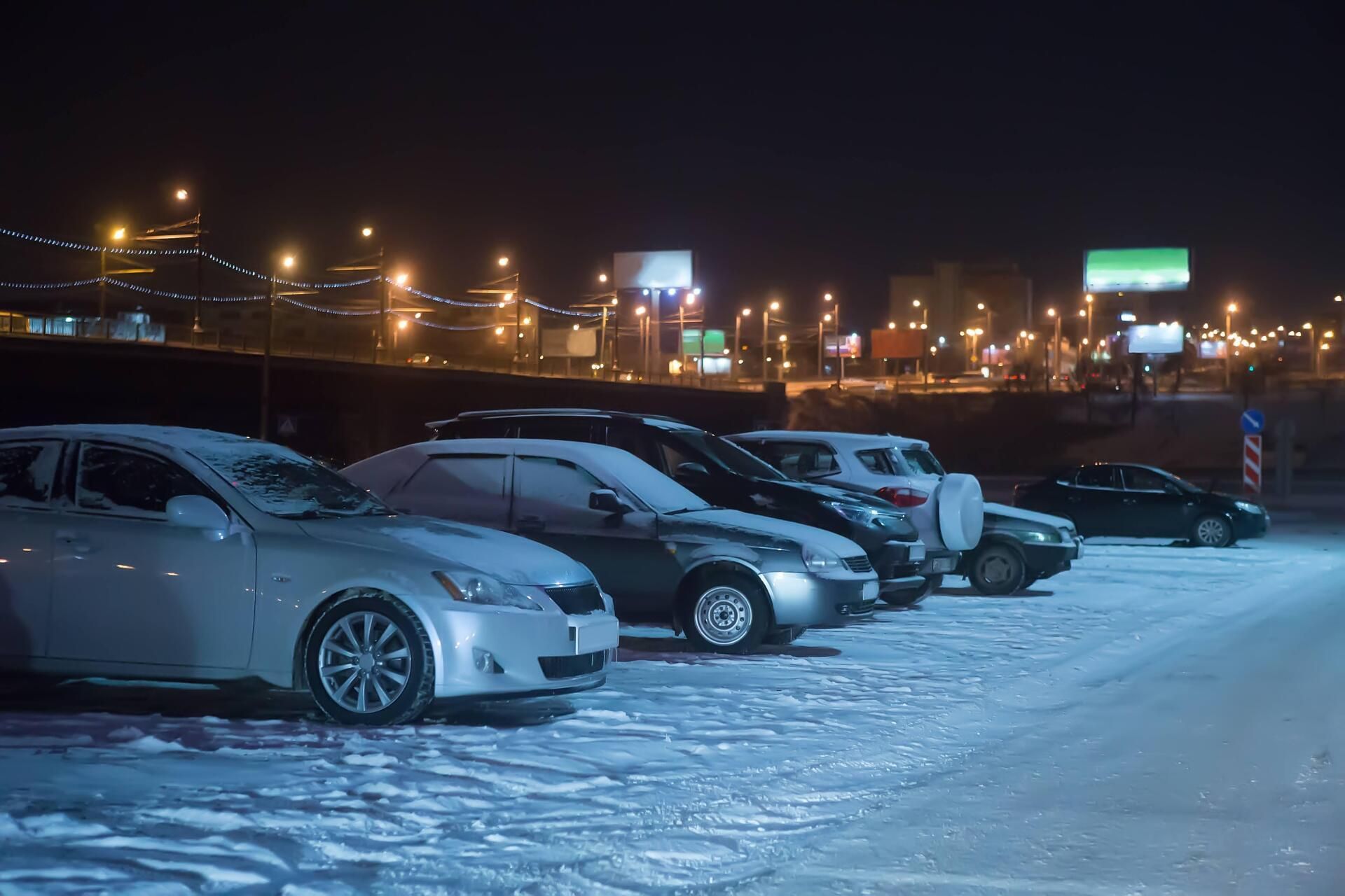 A row of cars are parked in a snowy parking lot at night.