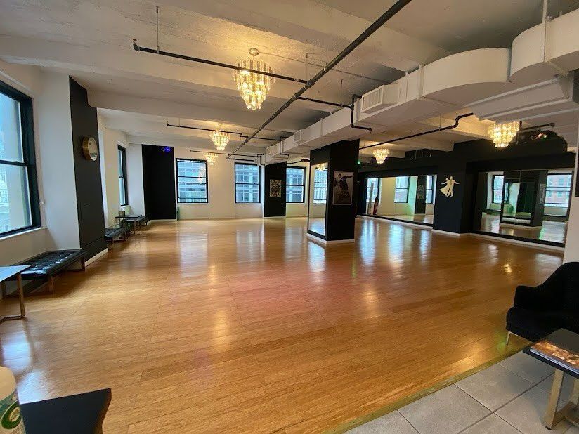 Empty dance studio with wood floor, mirrored walls, and large windows under ceiling lights.
