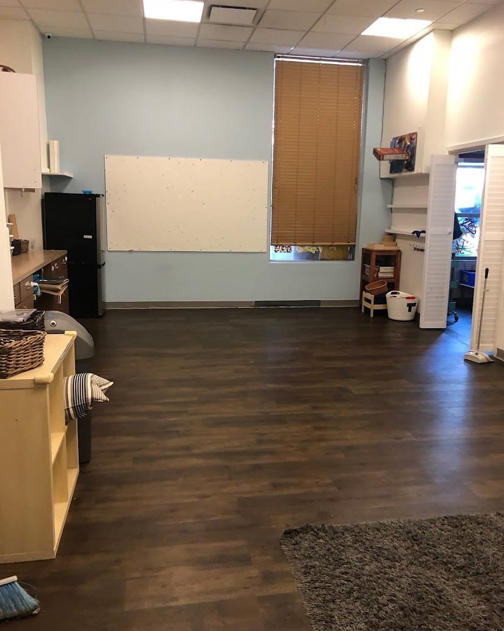 Empty office room with dark wood floor, white walls, window blinds, and shelves along the sides