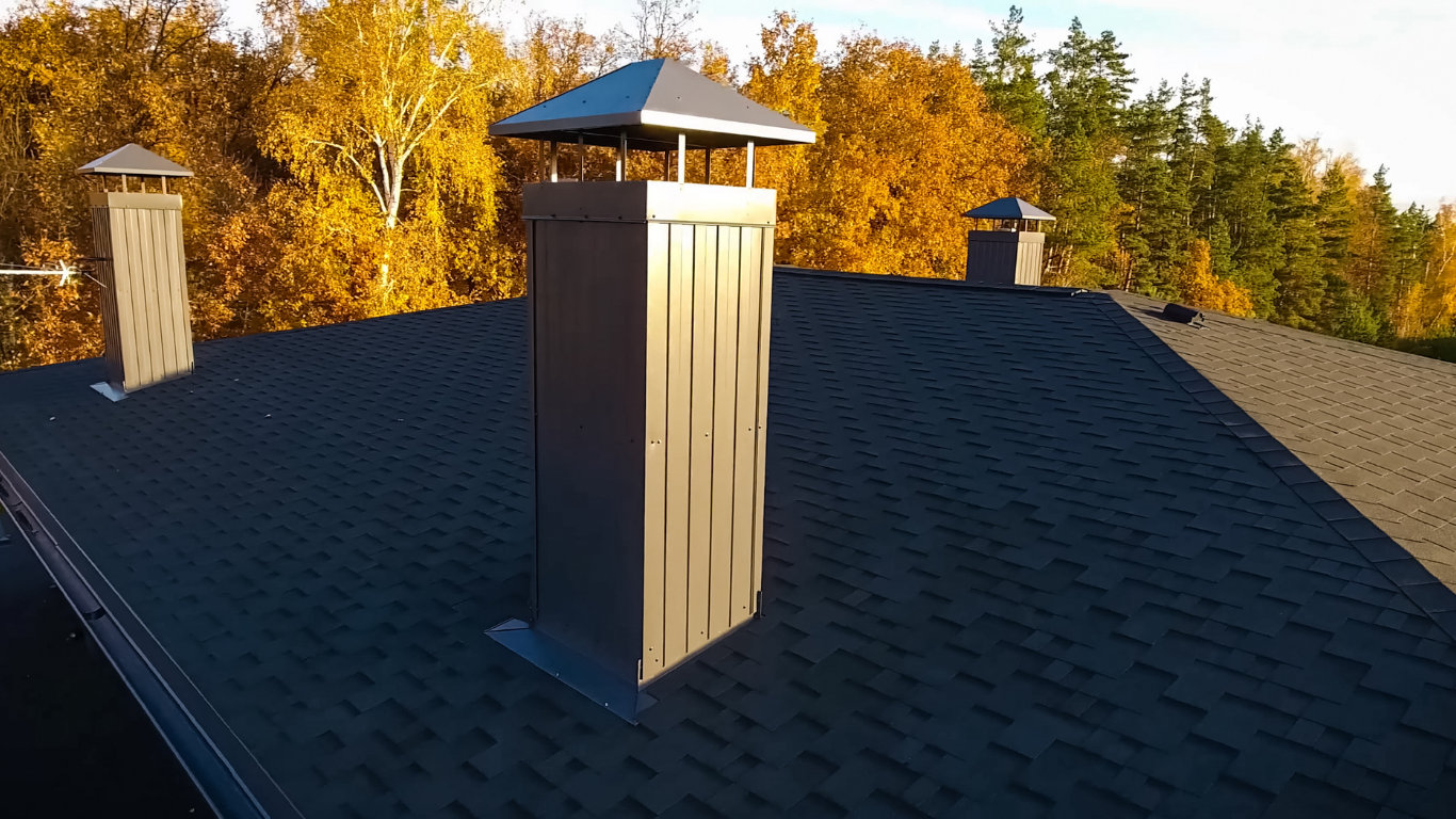 Rooftop with dark shingles and a silver chimney against autumn trees
