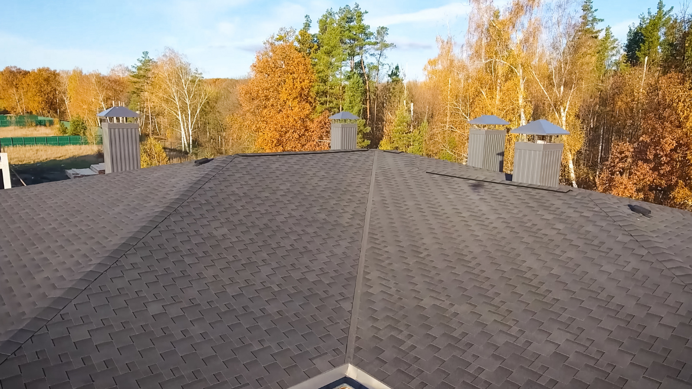 Rooftop with gray shingles and several chimneys, surrounded by autumn trees under a clear sky