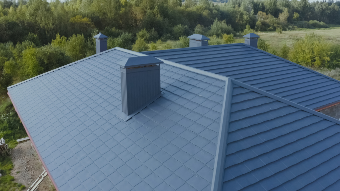 Blue metal roof with dormer vent, viewed from above against green trees.