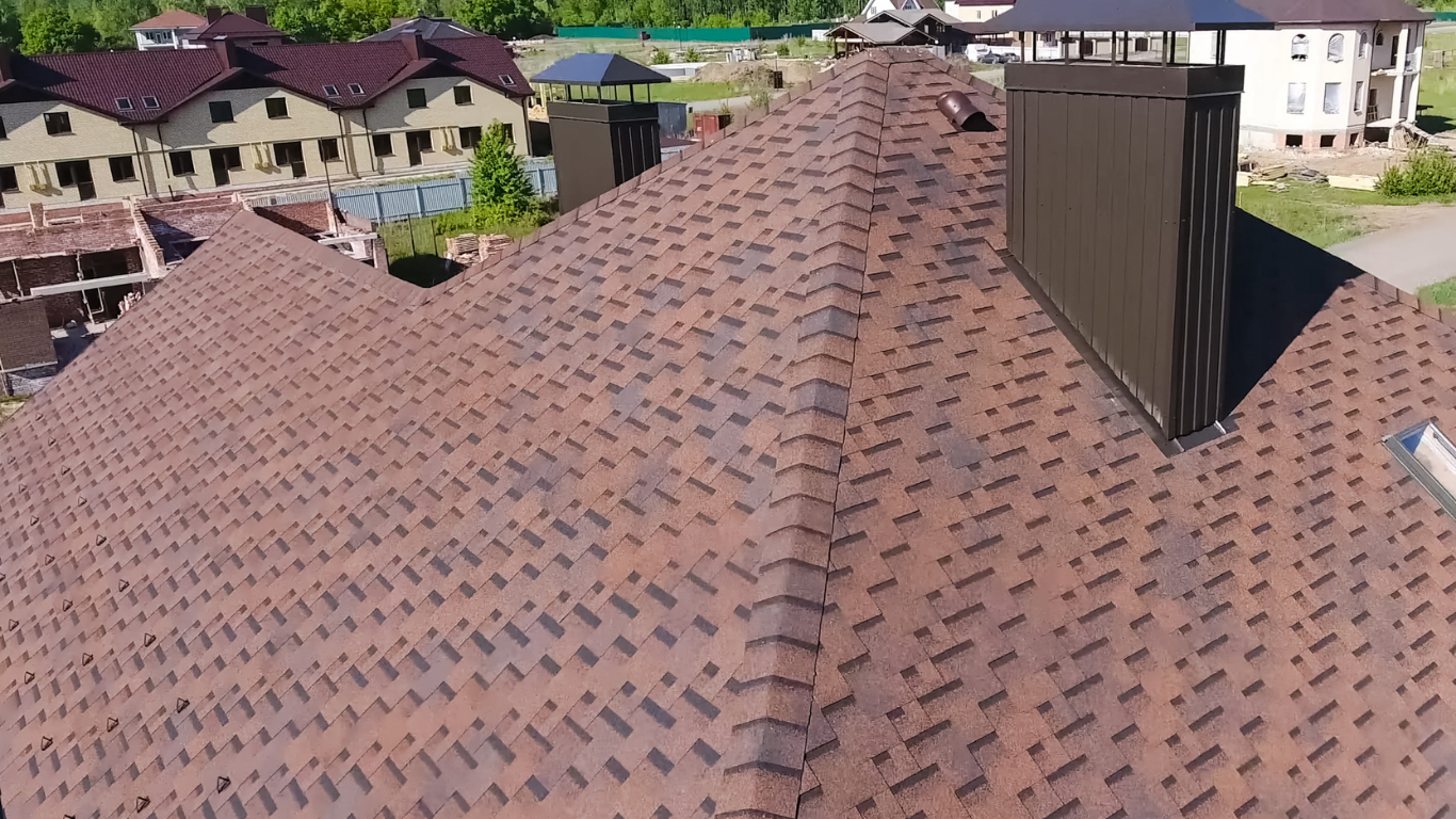 Aerial view of a brown shingle roof with a dark vent chimney in a suburban neighborhood.