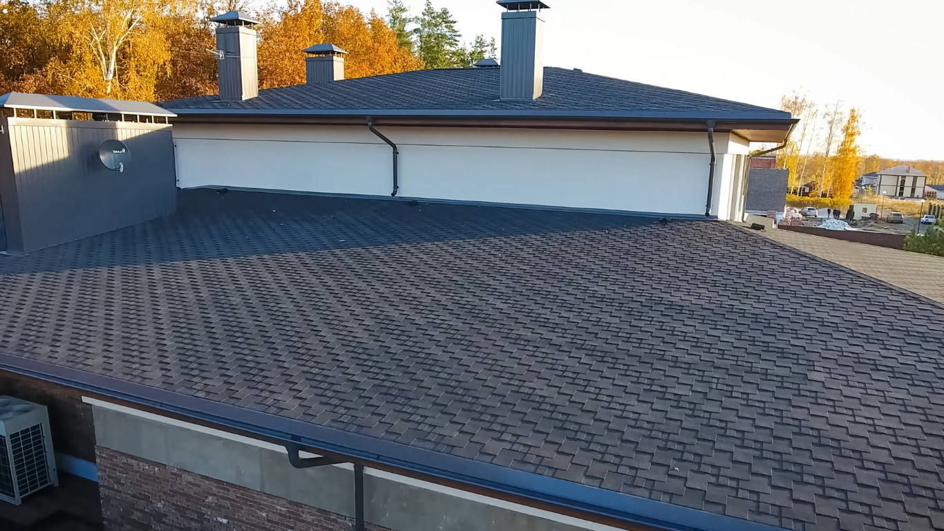 Rooftop with dark shingle tiles, white building walls, and chimneys at sunset.
