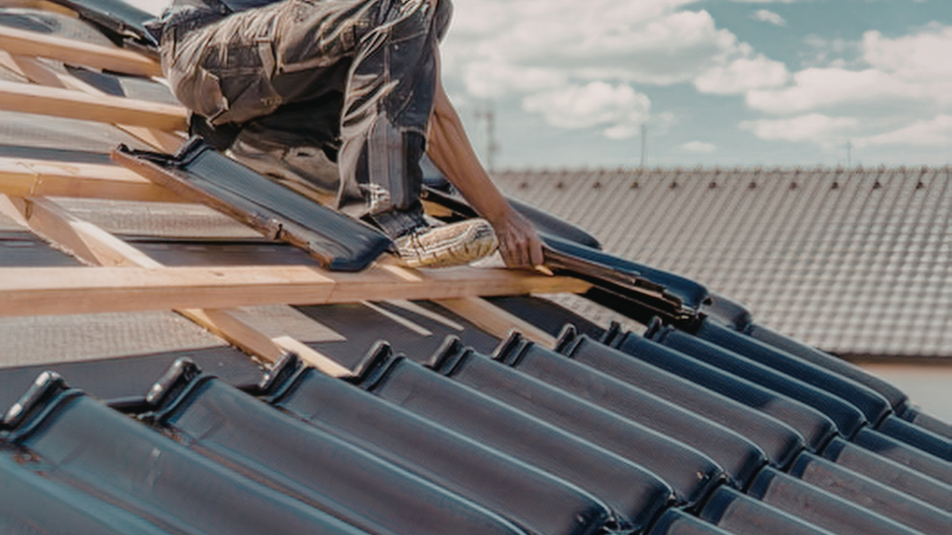 Worker installing dark blue roof tiles on a house roof with wooden framing visible