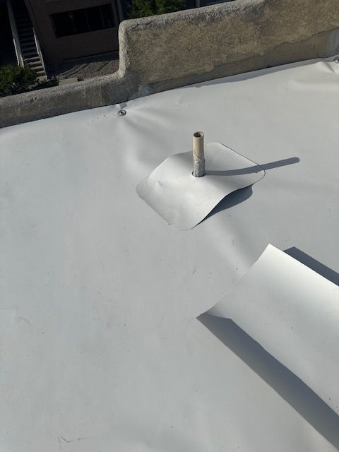 White rooftop membrane with a loose flap around a vent pipe near a concrete parapet.