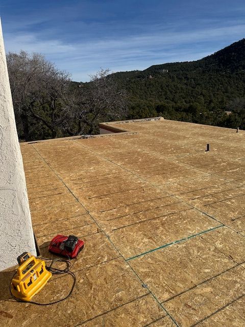 Rooftop under construction with plywood decking, yellow nailer, red tool bag, and forested hills beyond.