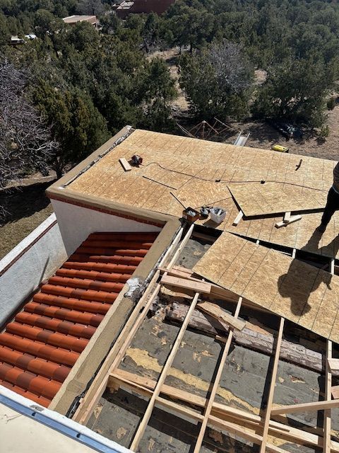 Roof under construction with exposed plywood, framing, and red tile roof beside trees
