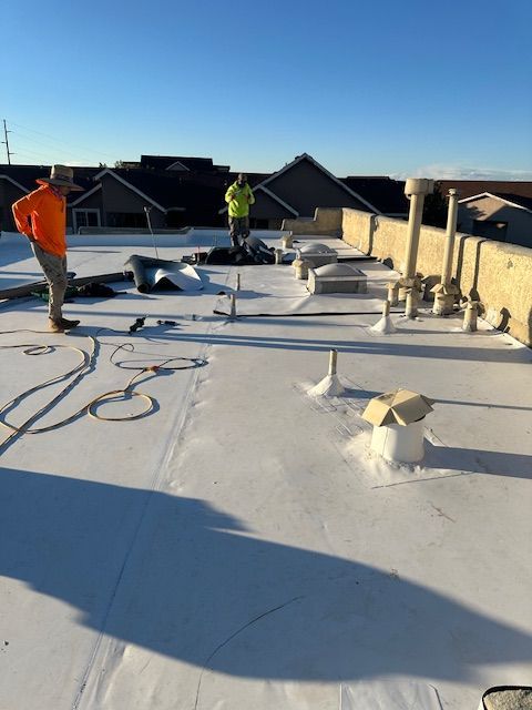 Workers on a flat roof installing white roofing membrane under a clear blue sky