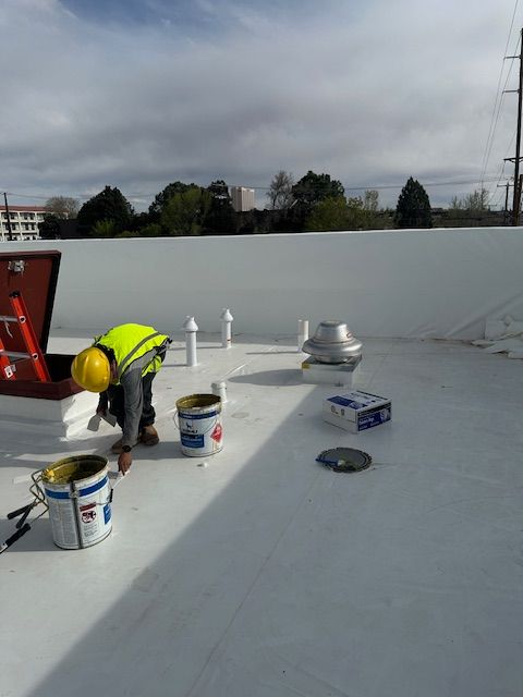 Worker in yellow hard hat applying coating on a flat rooftop with buckets and tools.