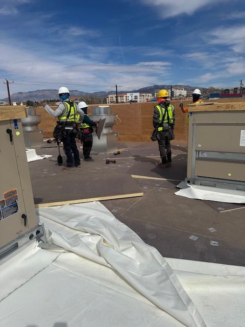 Workers in safety vests and helmets on a rooftop, handling materials near equipment under a blue sky.