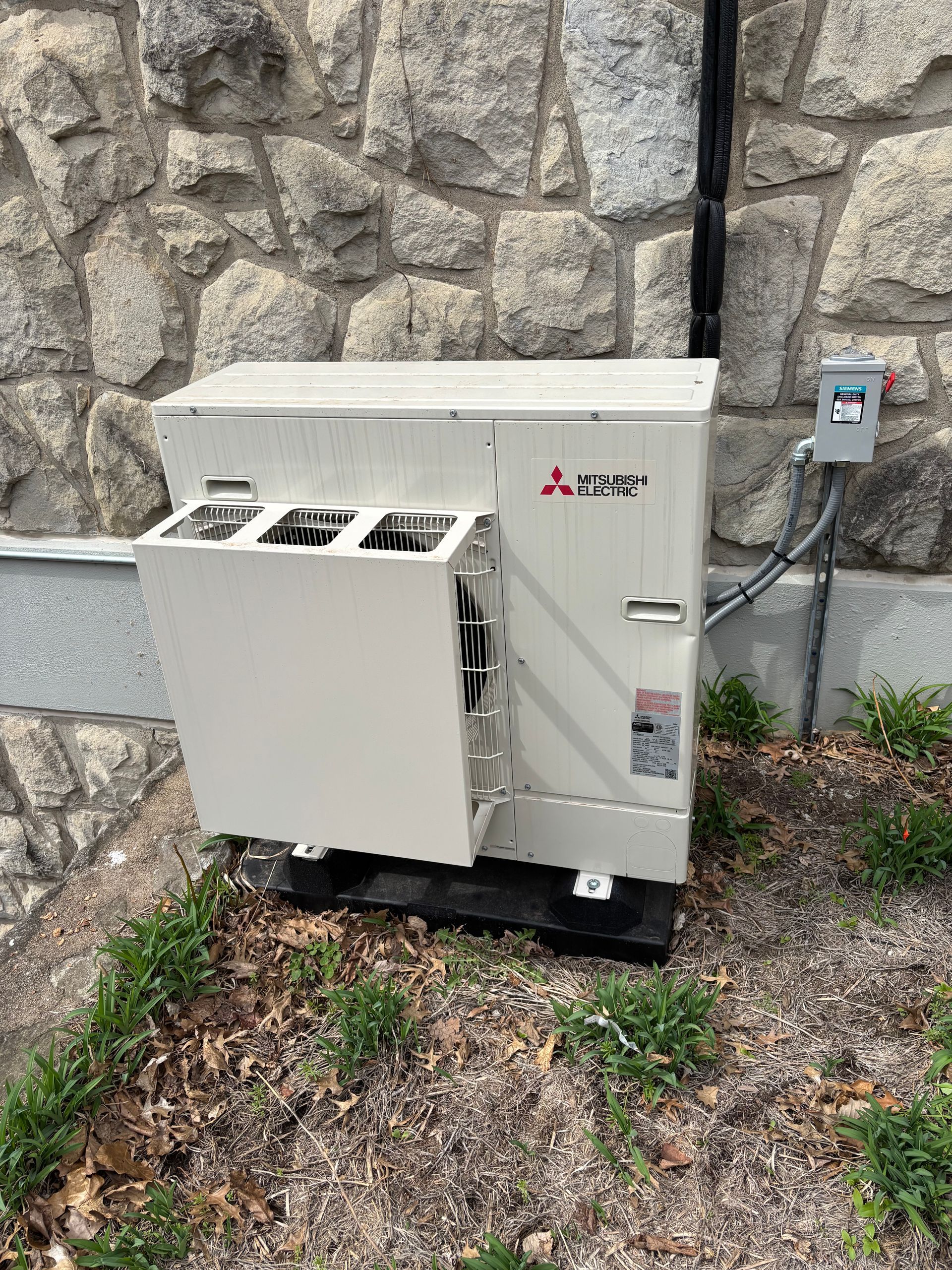 Mitsubishi outdoor HVAC unit against a stone wall with electrical components.