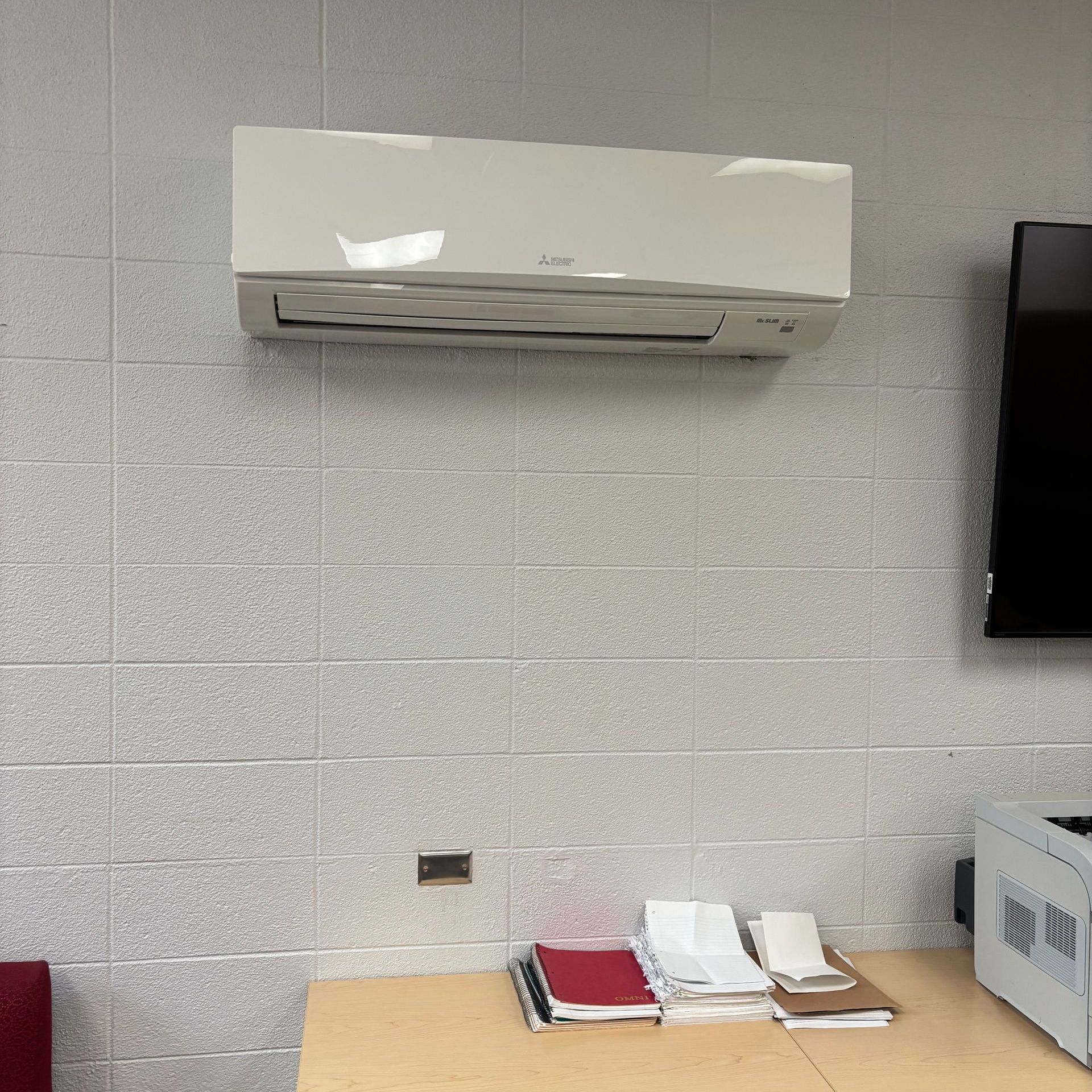 White air conditioner on a gray-tiled wall above a table with papers, printer, and red chair.