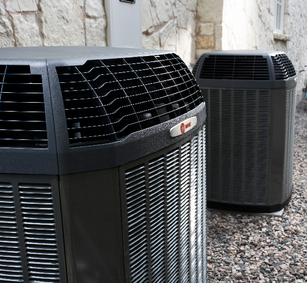 Two dark air conditioning units outside against a stone wall.